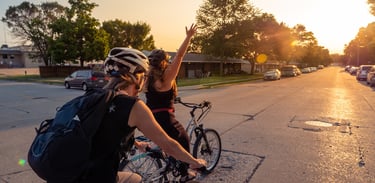 Image of two women bikers riding in the street, one with her arm stretched up in celebration