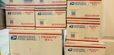 Stack of USPS Priority Mail boxes on a blue shipping cart ready for delivery or pickup.