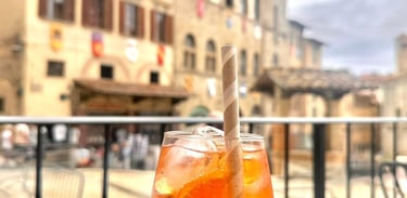Refreshing orange Aperol Spritz cocktail with a paper straw at an outdoor cafe in a historic Italian piazza.