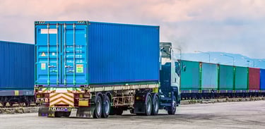 A logistics semi-truck carrying a blue shipping container near a freight train for intermodal transport.