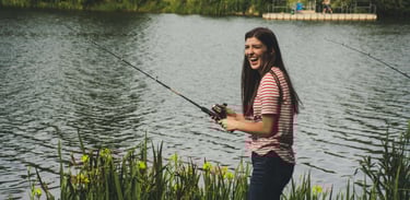 mulher morena com blusa listada pescando com vara de fibra e lago ao fundo.