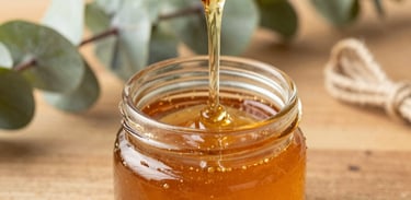 A close-up photograph of rich golden amber honey being poured from a dipper into a small glass favor jar. The jar rests on a warm wood brown surface surrounded by sage green eucalyptus leaves and twine. Gentle natural daylight.