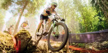 Professional mountain biker racing through a rocky forest trail during a cross-country competition.