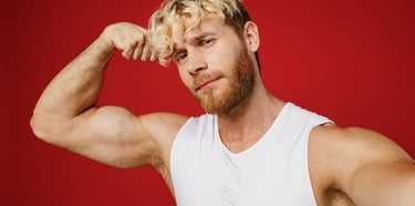 A blonde man with a beard flexes his bicep muscle while wearing a white tank top against a red background.