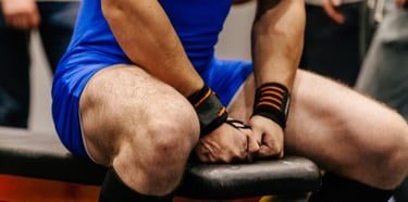 A powerlifter in a blue singlet wearing wrist wraps sits on a weight bench preparing for a lift.