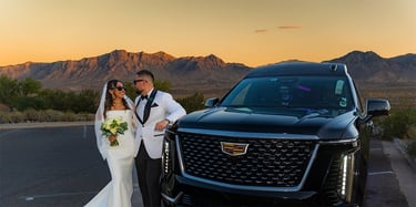 A bride and groom pose by a black Cadillac Escalade in the desert at sunset.