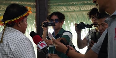 Indigenous leader in traditional headdress interviewed by journalists with microphones and cameras.