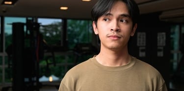 A young man in a tan sweatshirt standing in a modern gym with exercise equipment in the background.