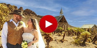 A smiling couple at a desert wedding in front of a rustic chapel and cactus plants.