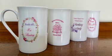 Four white bone china mugs with pink spicy romance book trope quotes on a wooden table.