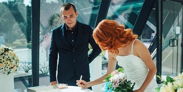 A redhead bride in a white wedding dress signs the marriage certificate during a modern indoor ceremony.