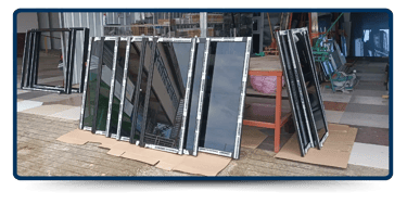 Several black-framed glass panels are neatly arranged on cardboard outside a workshop, with tools and materials