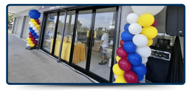 A festive entrance with colorful balloon arches beside glass doors, a table with a yellow cloth, and a person setting up