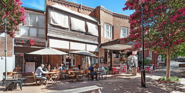 People dining at an outdoor sidewalk cafe with red brick buildings and blooming pink trees.