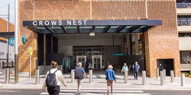 Commuters crossing the street toward the modern brick entrance of Crows Nest Metro station.