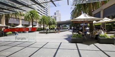 Sunny modern outdoor Forum Plaza with palm trees, glass awnings, and cafe seating in an urban shopping area.