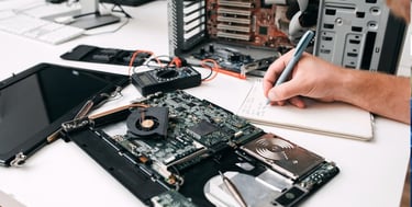 A technician repairing a laptop motherboard and taking notes for hardware diagnostics and computer maintenance.