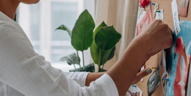 A creative woman pinning photos and inspiration to a cork mood board in a home office.