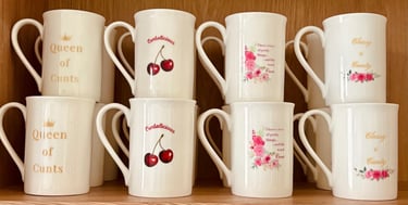 White bone china mugs with floral designs and bold profane slogans on a wooden shelf.