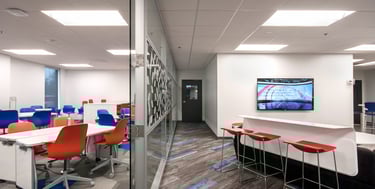 Modern office collaboration space with colorful seating, glass partitions, and a wall-mounted TV.