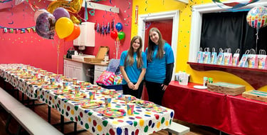 Two party hosts standing in a colorful kids birthday party room with decorated tables and balloons.