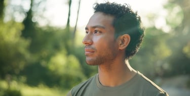 A profile of a young man with curly hair standing in a sunlit park with lens flare and greenery.