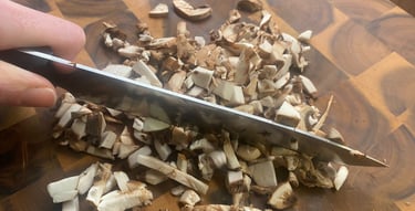 Close-up of a hand chopping fresh white button mushrooms on a wooden cutting board with a silver chef's knife, preparing