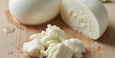 Fresh mozzarella cheese: a whole ball, a cut half, and torn pieces, with basil leaves on a wooden board.