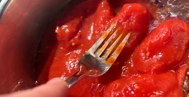 A metal fork mashing whole peeled San Marzano tomatoes in a stainless steel pot for a homemade pasta sauce.