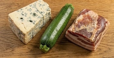 Fresh wedge of blue cheese, a whole green zucchini, and cured pancetta meat on a wooden table.