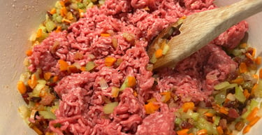 Browning raw ground beef with sautéed carrots, onions, and celery in a white pot for bolognese sauce.