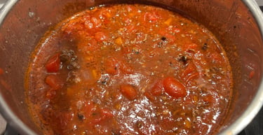 Homemade bolognese meat sauce with chunks of tomato simmering in a stainless steel pot on a stove.