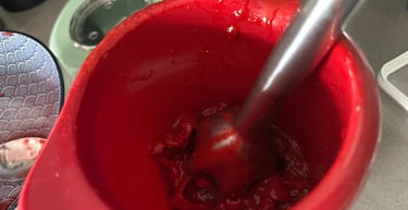 Stainless steel hand immersion blender pureeing red fruit in a red mixing bowl for a smoothie.