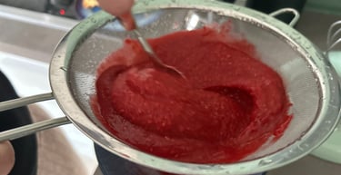 A person using a metal mesh strainer to filter fresh raspberry puree for a seedless fruit sauce.