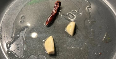 Fresh garlic cloves and a dried red chili pepper sizzling in olive oil inside a non-stick frying pan.