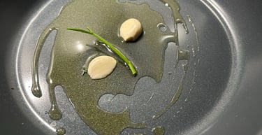 Peeled garlic cloves and a herb stem sizzling in olive oil inside a non-stick frying pan.