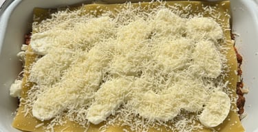 Homemade lasagna layers topped with fresh mozzarella slices and shredded parmesan cheese in a white baking dish.
