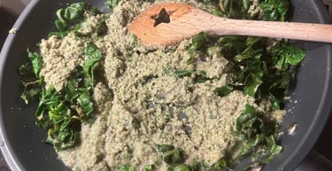 Sautéed green leafy vegetables and creamy pesto sauce cooking in a black frying pan with a wooden spoon.