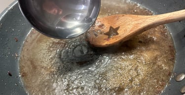 A metal ladle pouring water into a bubbling pan of broth for deglazing with a wooden spoon.