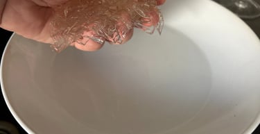 Hand holding softened gelatin sheets over a white bowl of water for a recipe.