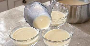 A metal ladle pouring creamy vanilla custard into glass dessert bowls in a kitchen.