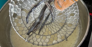 Removing infused vanilla bean pods from a simmering pot of custard with a metal spider strainer.