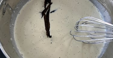 Whisking fresh vanilla bean pods into a creamy white custard base in a stainless steel pot.