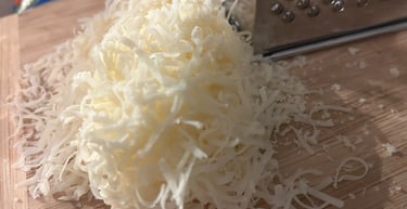 Freshly shredded white cheddar cheese piled on a wooden cutting board next to a metal box grater.