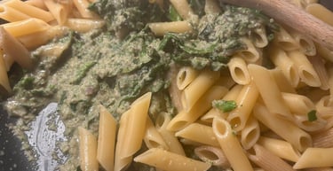 Cooked penne pasta being tossed with creamy green spinach pesto sauce in a large skillet.