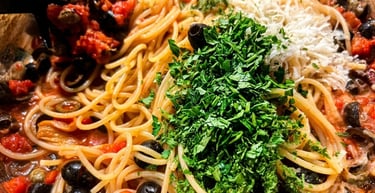 Spaghetti puttanesca with black olives, tomatoes, fresh parsley, and parmesan cheese in a skillet.