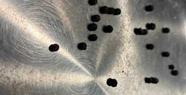 Whole black peppercorns scattered on a textured stainless steel surface with bright reflections.