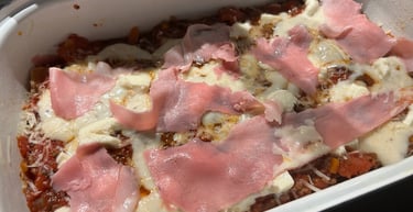 Homemade lasagna in a white baking dish topped with tomato sauce, melted mozzarella, and thin ham slices.