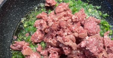 Raw ground Italian sausage cooking in a skillet with chopped green onions and olive oil.