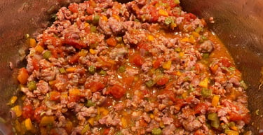 Ground beef simmering with diced tomatoes, carrots, and celery in a metal pot for meat sauce.